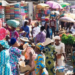 Busy outdoor market with people in colorful traditional clothing, surrounded by stalls selling produce and textiles under umbrellas.